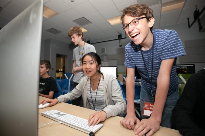 a high school student leans over a computer and laughs as he watches another student play their original video game created at the 2019 SDCTx Game Design Summer Institute