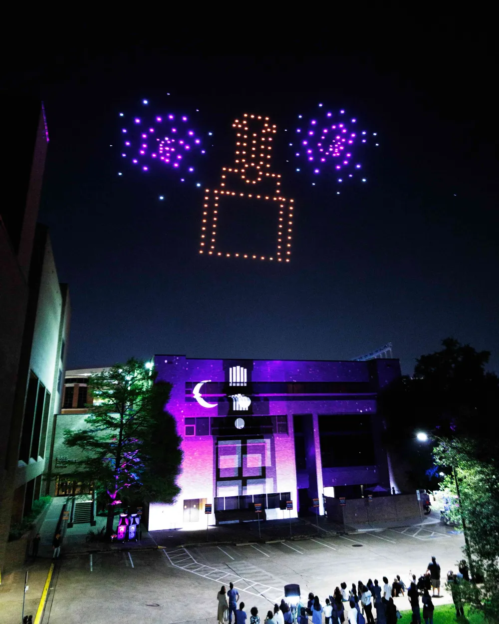 A drone light show forms the shape of a robot figure flanked by glowing orbs in the night sky above a purple-lit building on the University of Texas at Austin campus, as a crowd watches from below.