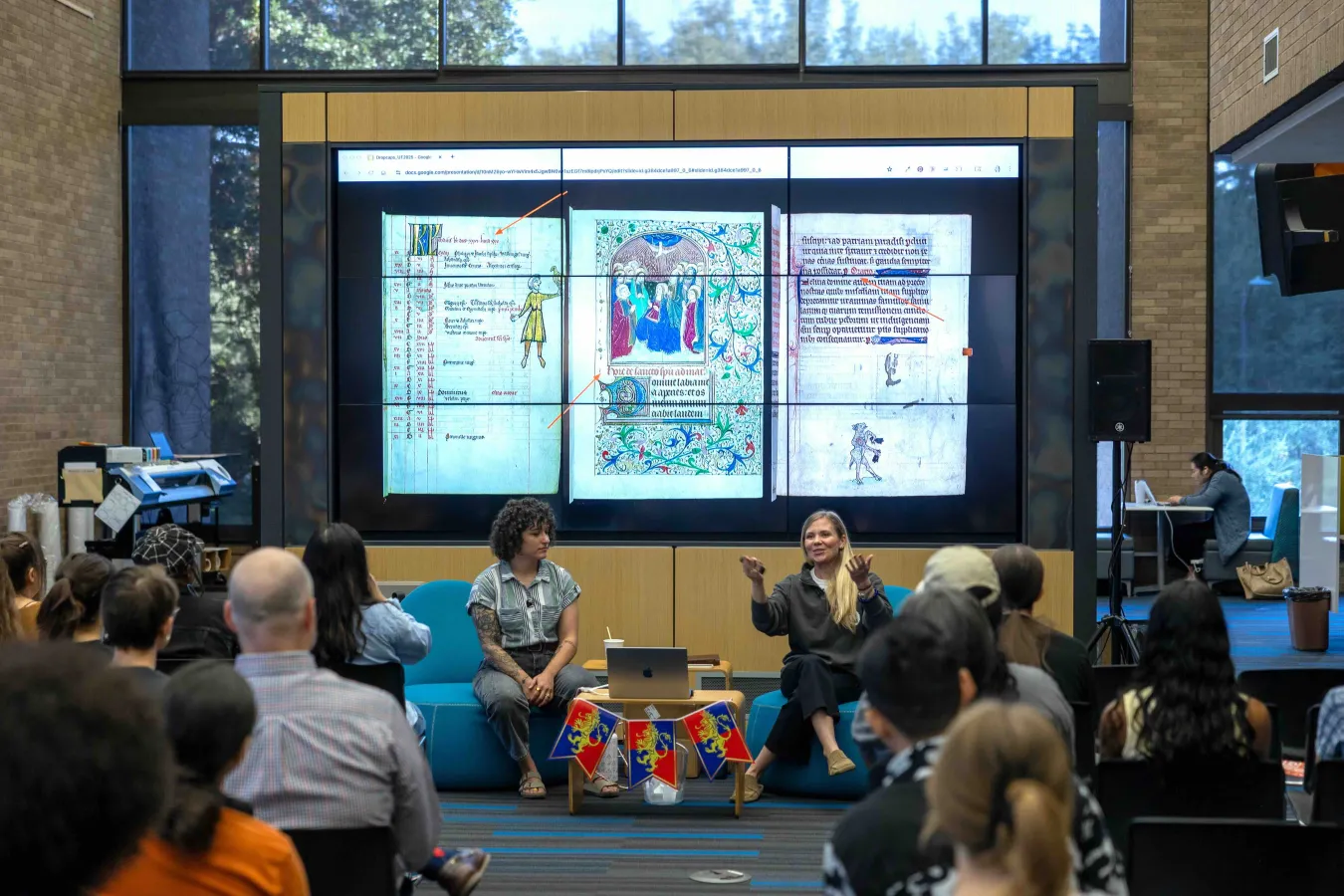 Two speakers lead a presentation in front of a seated audience, with a large screen behind them displaying illuminated medieval manuscripts and colorful annotations.