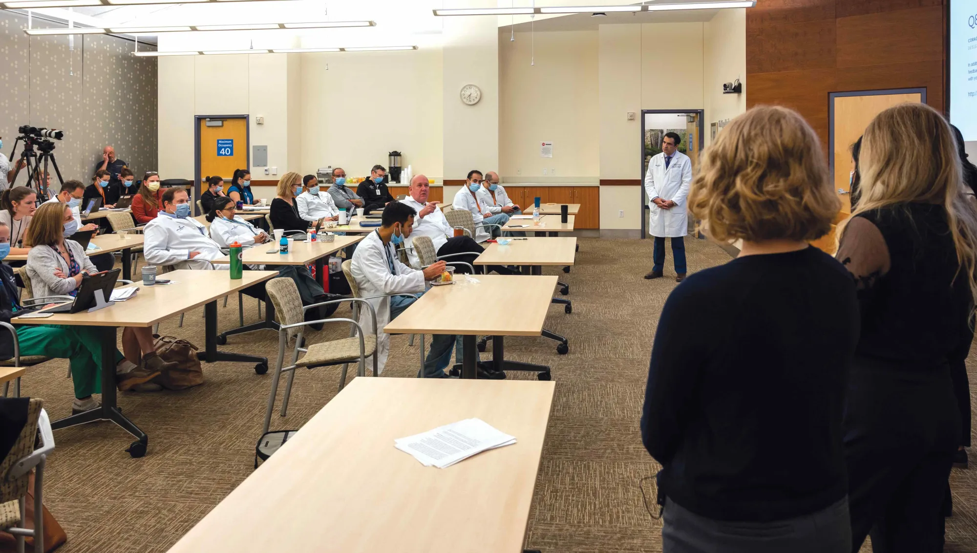 A packed conference room filled with medical professionals seated at long tables, attentively listening to a presentation. Many attendees wear white lab coats and surgical masks, indicating a healthcare setting. Two presenters stand at the front facing the audience, while a third person in a lab coat stands near the screen. The space is well-lit, with cameras positioned in the back capturing the event.