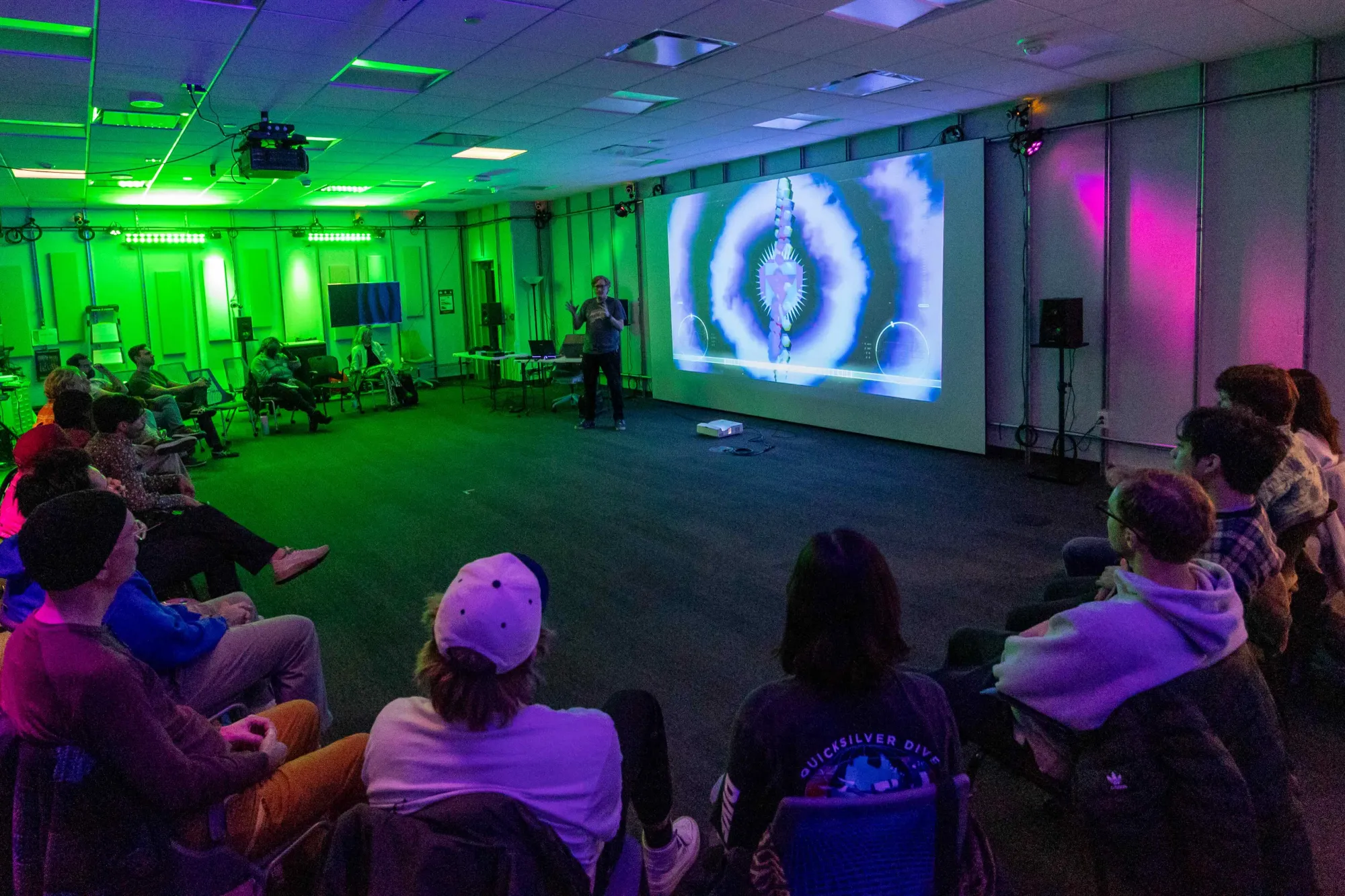A presenter speaks to an audience seated in a dark room illuminated by green and purple lights. A vibrant projection featuring abstract visuals is displayed on a large screen behind the speaker.