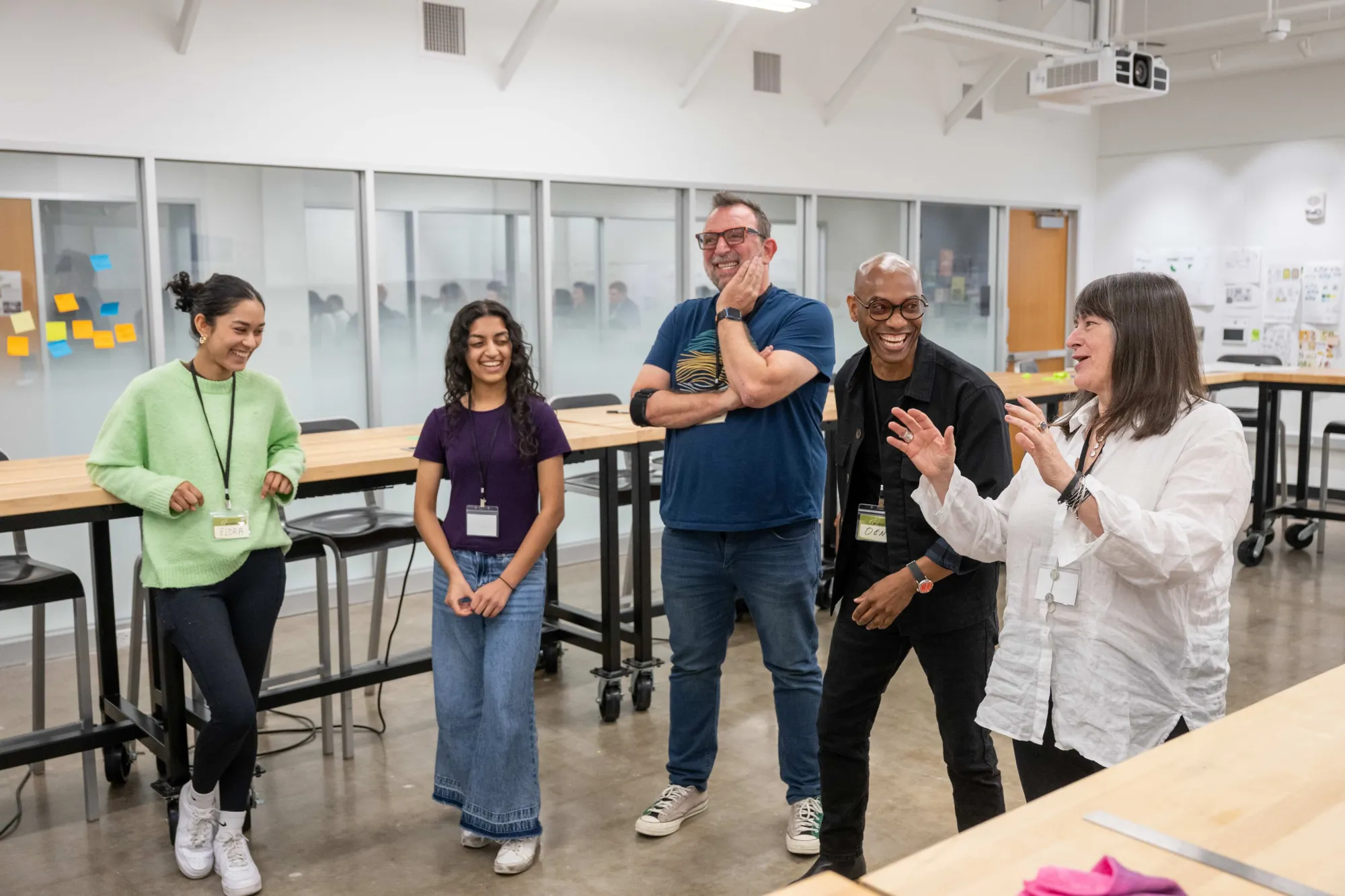 A group of five people—two students and three instructors—stand together in a design studio classroom, smiling and laughing during a collaborative activity. Large wooden tables and glass-walled meeting rooms are visible in the background.