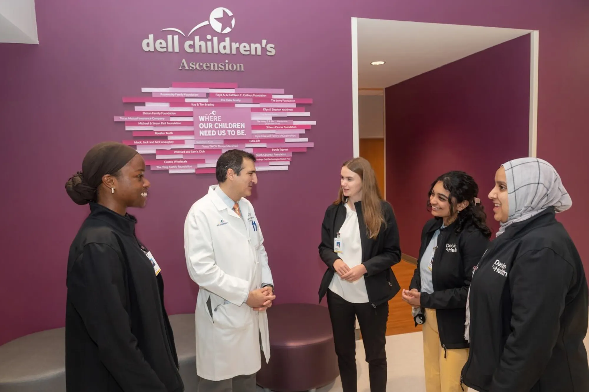 Five medical professionals stand in conversation in front of a purple wall at Dell Children’s Medical Center. The group includes four women and one man, with the man wearing a white lab coat and the others in black jackets labeled “Design for Health.” A sign on the wall behind them features donor names and the phrase “Where our children need us to be.”