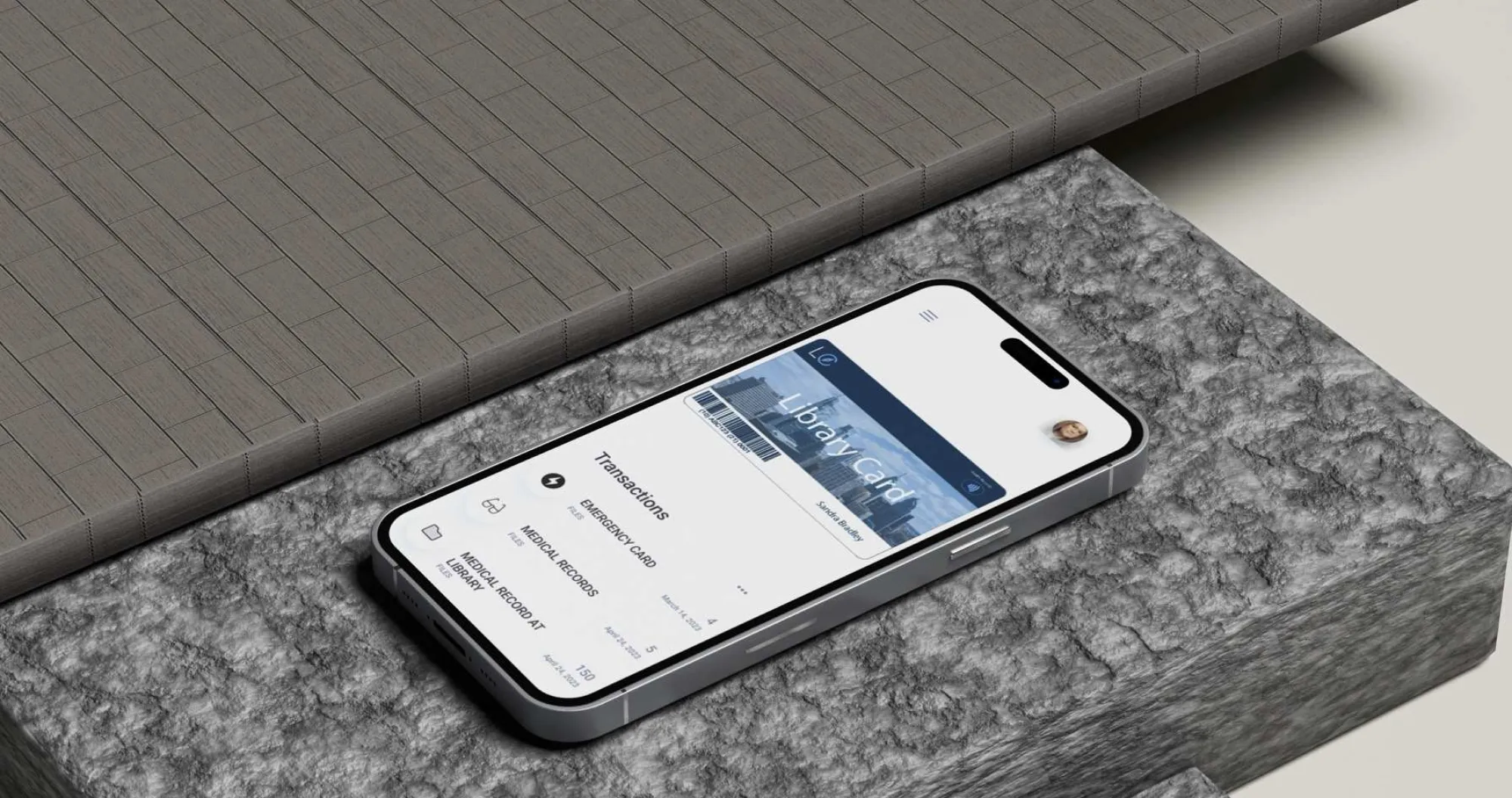 A smartphone resting on a stone-textured surface displays a digital version of a "Library Card" featuring a cityscape and barcode. The app interface shows recent transactions under the “Emergency Card” and “Medical Records” sections. A user profile icon appears in the upper-right corner, and the name "Sandra Bradley" is visible on the digital card. The screen design suggests library cards integrated with health and emergency record management.