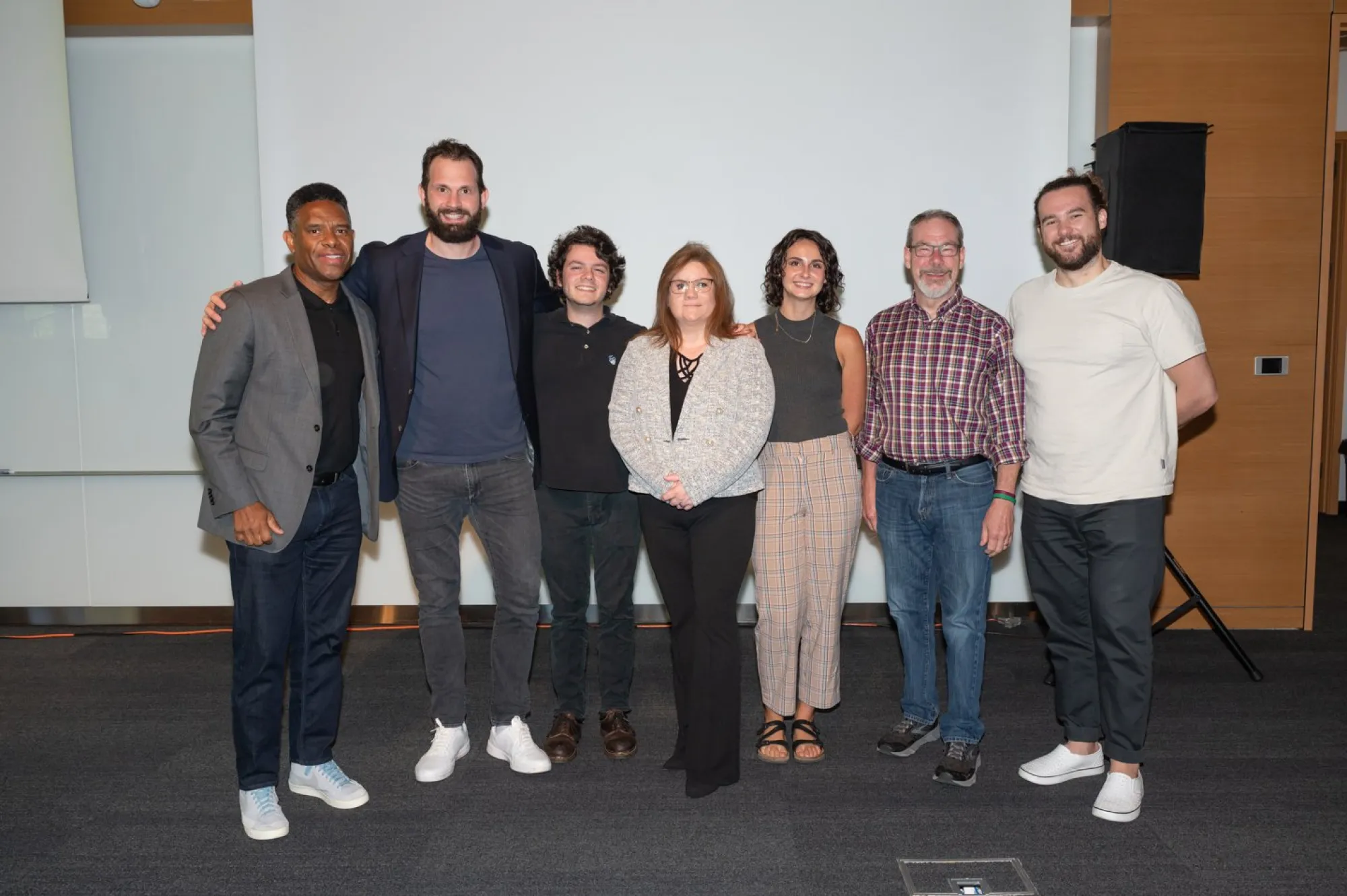 Seven people stand side by side in front of a projection screen, smiling for a group photo in a professional setting. They are dressed in business casual attire and appear to be a mix of presenters, faculty, and team members at a design or tech event. The environment suggests a conference room or lecture space with wood paneling and a visible speaker in the background.