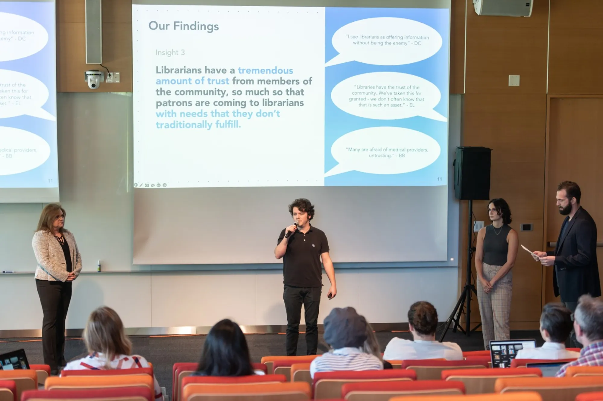 Four presenters stand at the front of a lecture hall speaking to an audience, with a large slide projected behind them. The slide is titled “Our Findings” and highlights Insight 3: Librarians have a tremendous amount of trust from members of the community, so much so that patrons are coming to librarians with needs that they don’t traditionally fulfill. Three speech bubbles on the right quote interviewees discussing librarians' trustworthiness and their role in healthcare information. Audience members are s