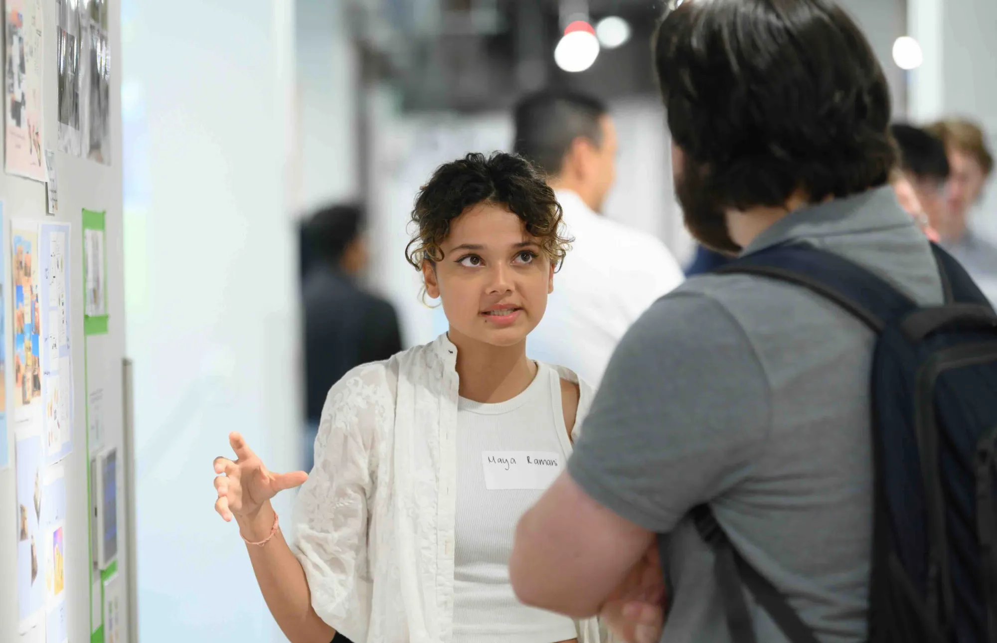 A student wearing a name tag that reads “Maya Lamari” presents her design work to an attendee at an exhibition. She gestures with her hands while speaking, standing beside a wall covered in printed visual materials. The background is softly blurred, showing other people engaged in conversation.