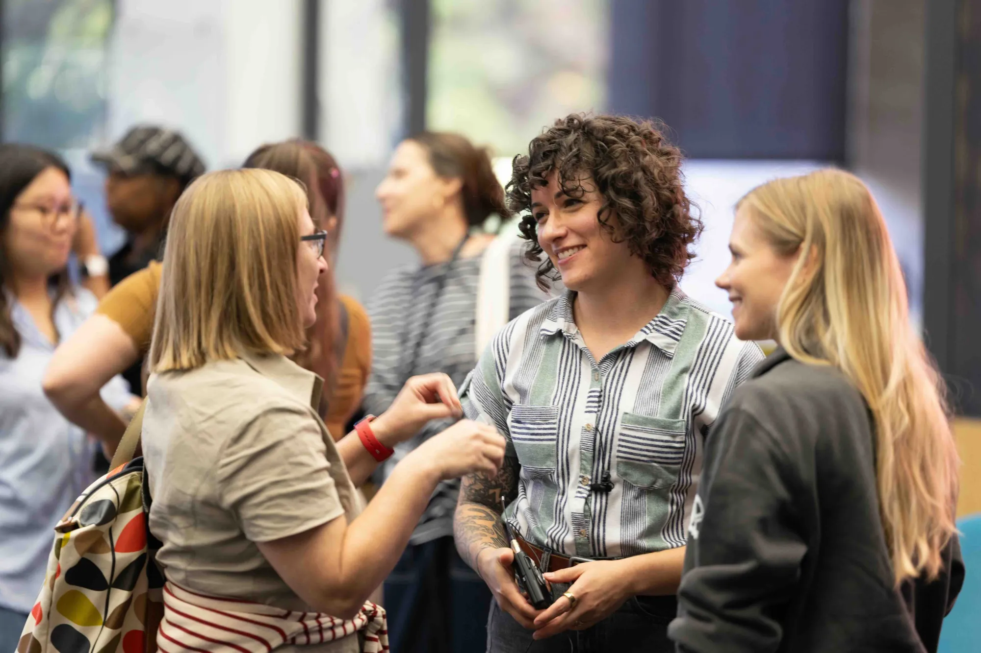 Three people smile and chat during a post-event gathering, while others engage in conversation in the blurred background of the room.