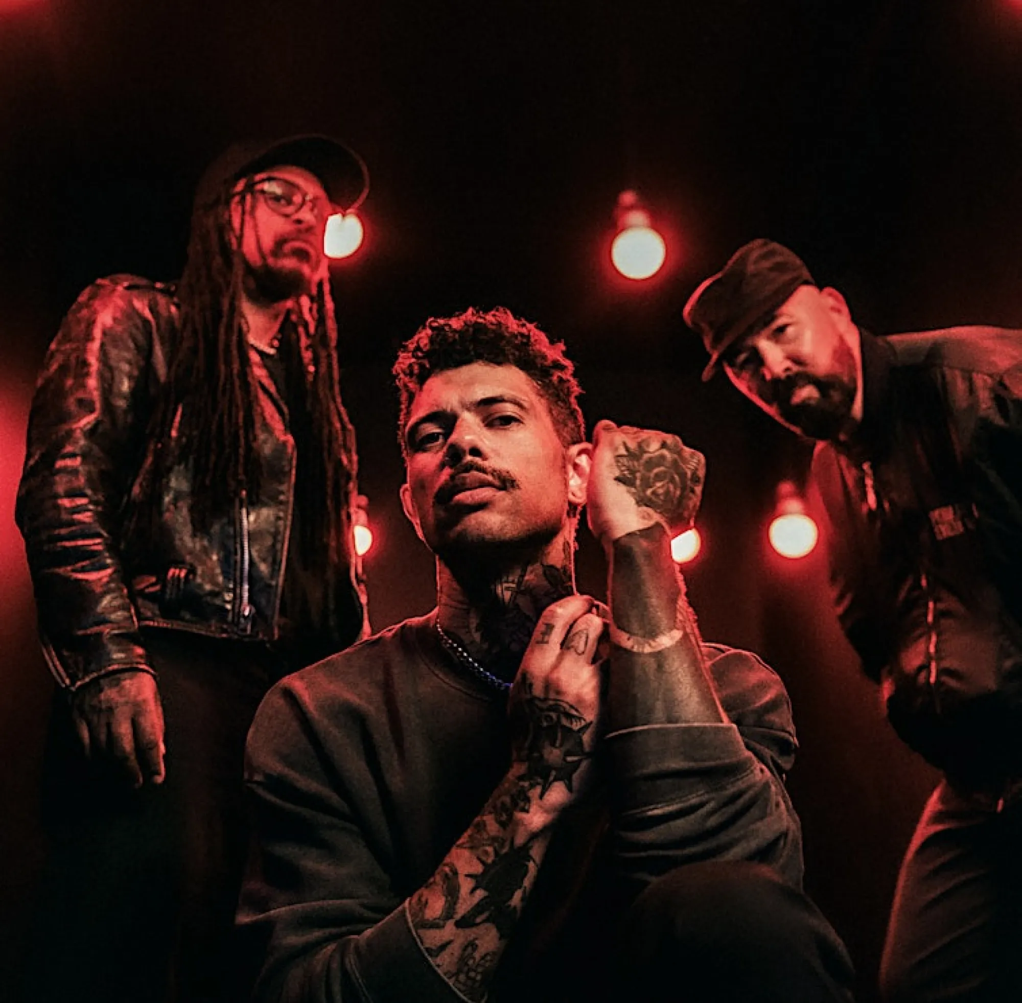 Dramatic portrait of the three members of Urban Heat, lit by red hanging lights. The central figure crouches with tattoos visible on his arms and hand, while the two other band members stand behind him, wearing dark clothing and looking into the camera.