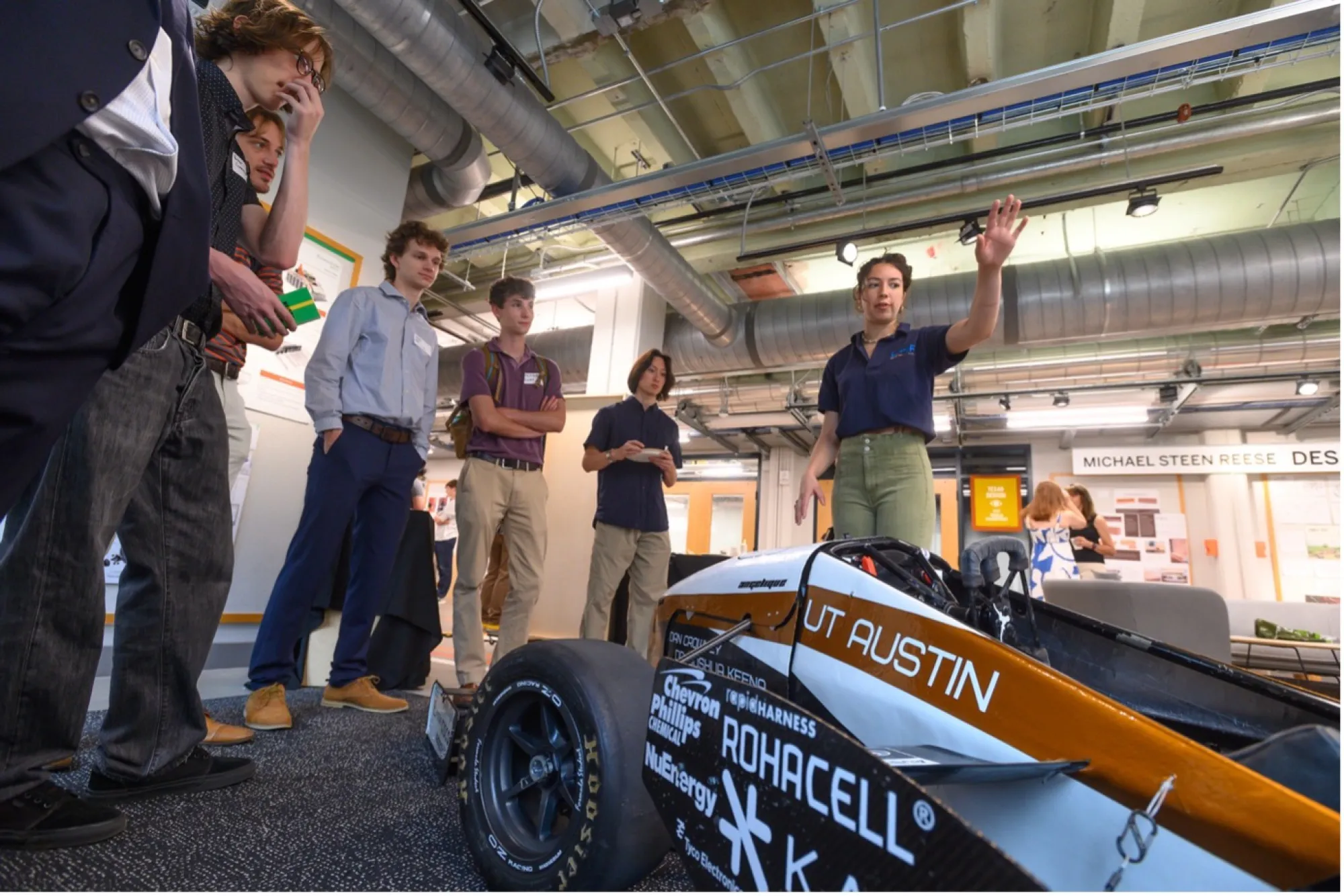 A UT Austin student gestures while presenting a Formula SAE race car to a group of prospective students in a design studio. The car is marked with sponsor logos and “UT Austin” in burnt orange text.