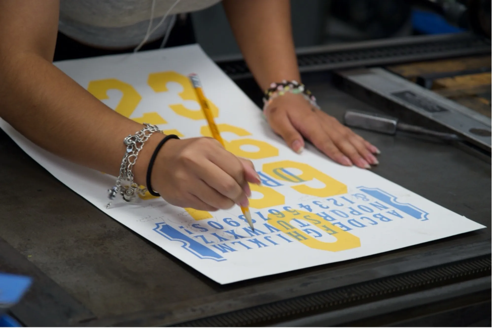 A student sketches details onto a print featuring large yellow numbers and blue letters. Their hands rest on the paper as they work at a letterpress station.
