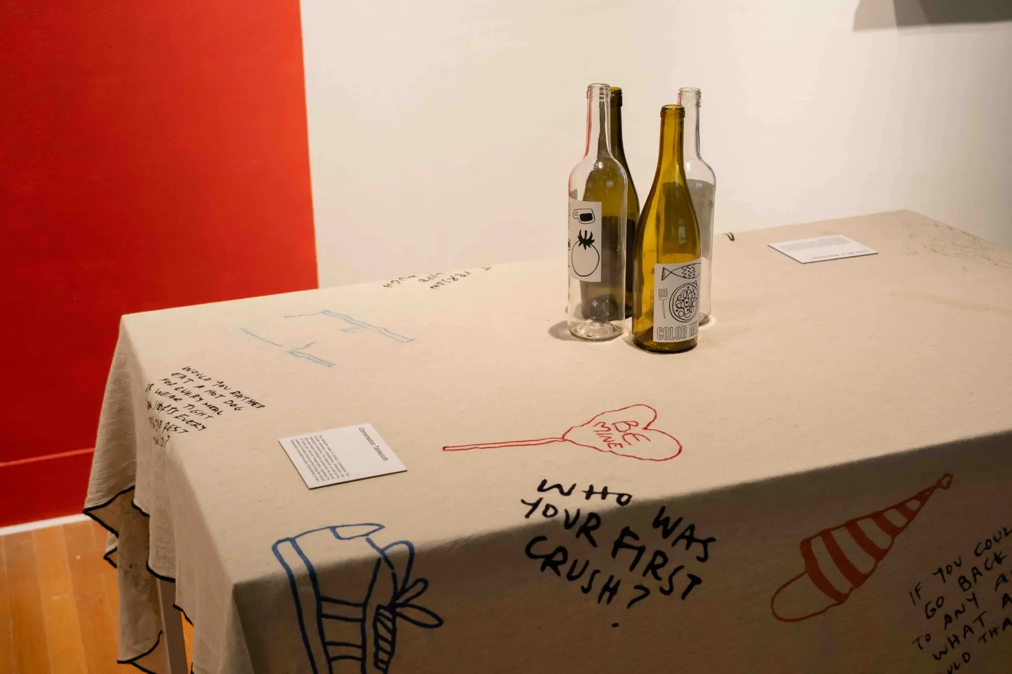 A linen-covered table features playful, hand-drawn illustrations and handwritten questions like “Who was your first crush?” surrounded by doodles of sneakers, party hats, and conversation hearts. Three glass bottles with custom illustrated labels sit at the center, inviting viewers into an interactive, conversational installation.