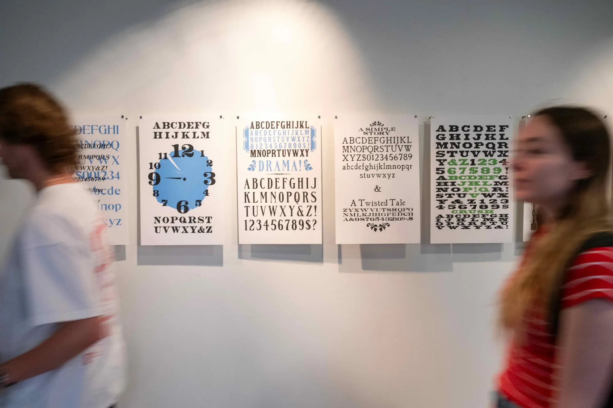 Two students walk past a gallery wall displaying a series of typographic posters, each featuring a distinct typeface arranged in creative compositions. One poster mimics a clock face with numbers replacing hour markers, while others explore font pairings, stylistic sets, and decorative elements, emphasizing expressive storytelling through type design.