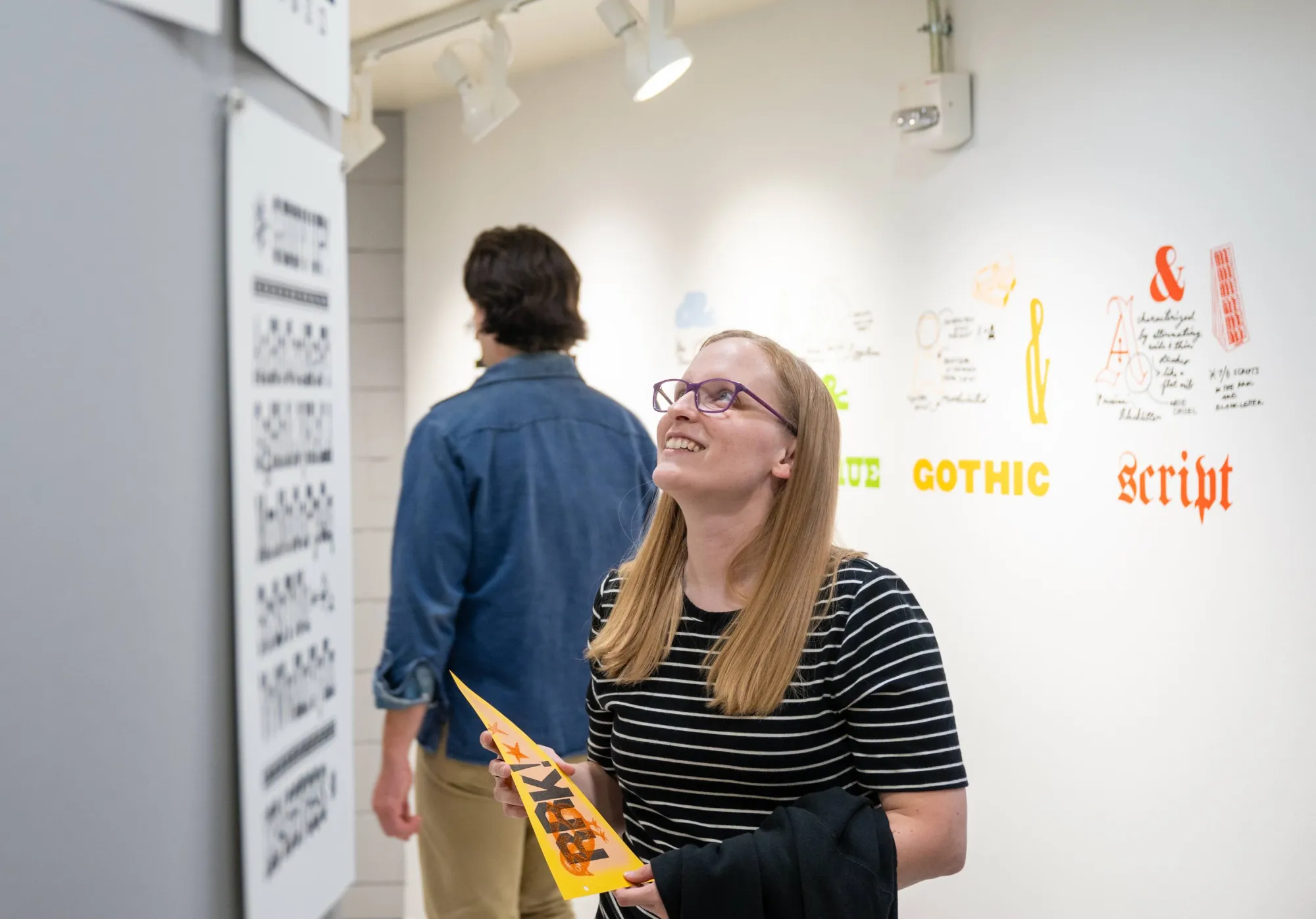 A smiling woman wearing glasses and holding a bright orange RRK pamphlet admires letterpress prints on display at the Rob Roy Kelly American Wood Type Collection exhibition. Behind her, another visitor browses the installation, and colorful type classifications like “Gothic” and “Script” are painted on the gallery wall.