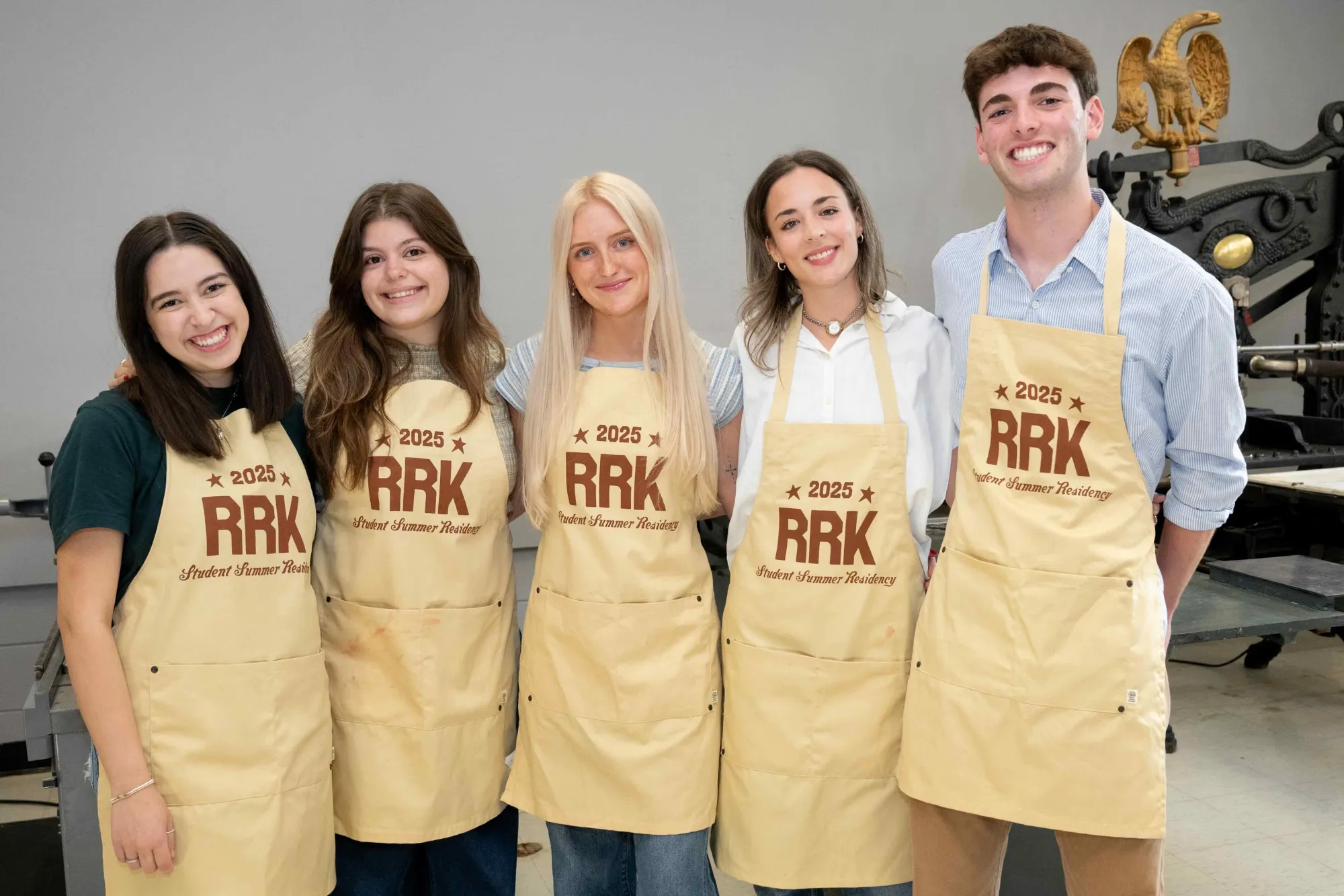 Five students smile and stand side-by-side wearing matching aprons printed with “2025 RRK Student Summer Residency.” Behind them, a vintage letterpress machine and eagle sculpture hint at the historic design tools used during the residency, part of the Rob Roy Kelly American Wood Type Collection.
