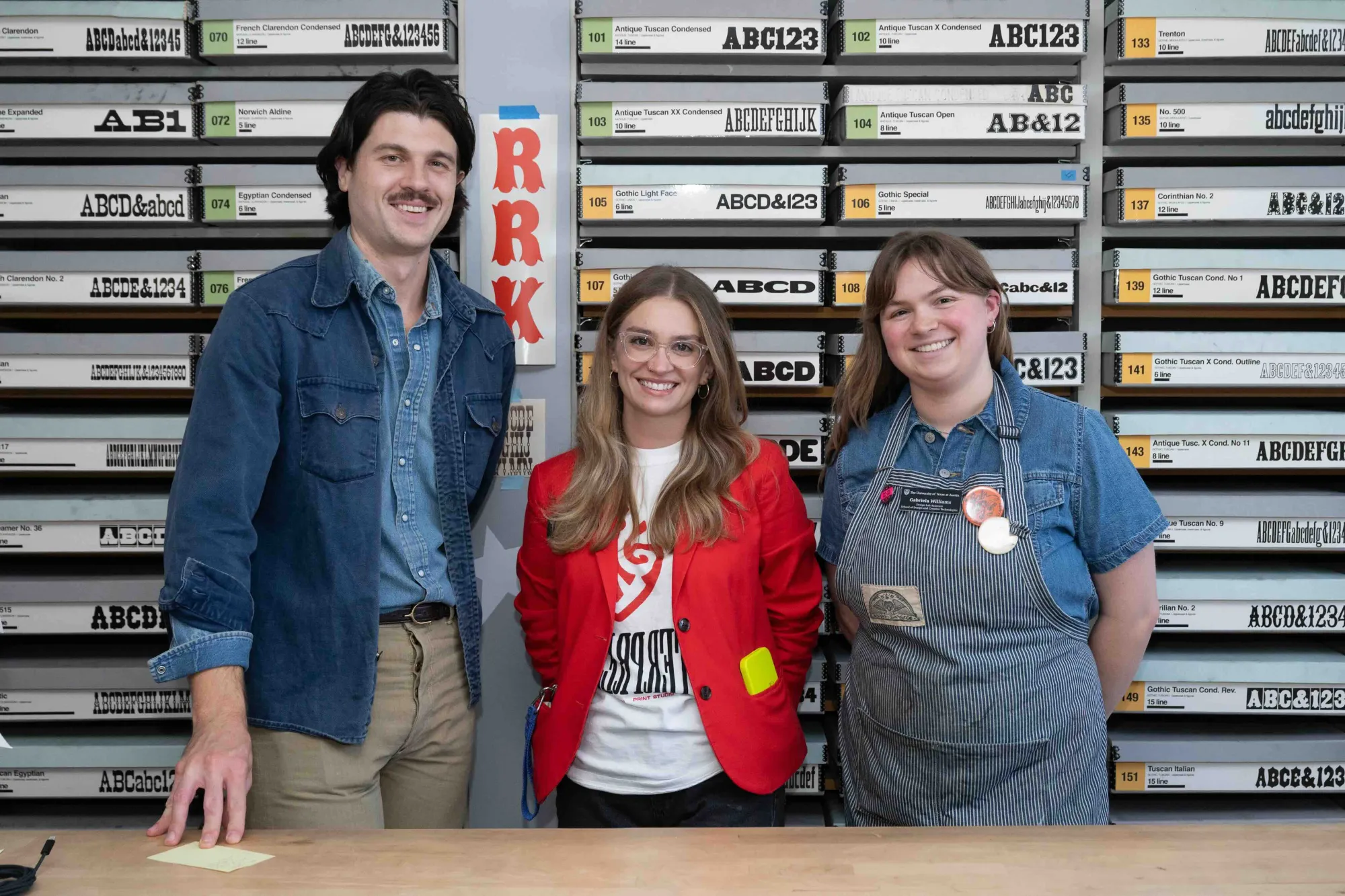 Three people stand smiling in front of a wall of labeled wood type specimen drawers, organized by typeface. A red vertical “RRK” banner visually anchors the scene, highlighting the Rob Roy Kelly American Wood Type Collection. Their outfits—denim, a bright red blazer, and a striped apron—capture a mix of vintage craft and contemporary design energy.