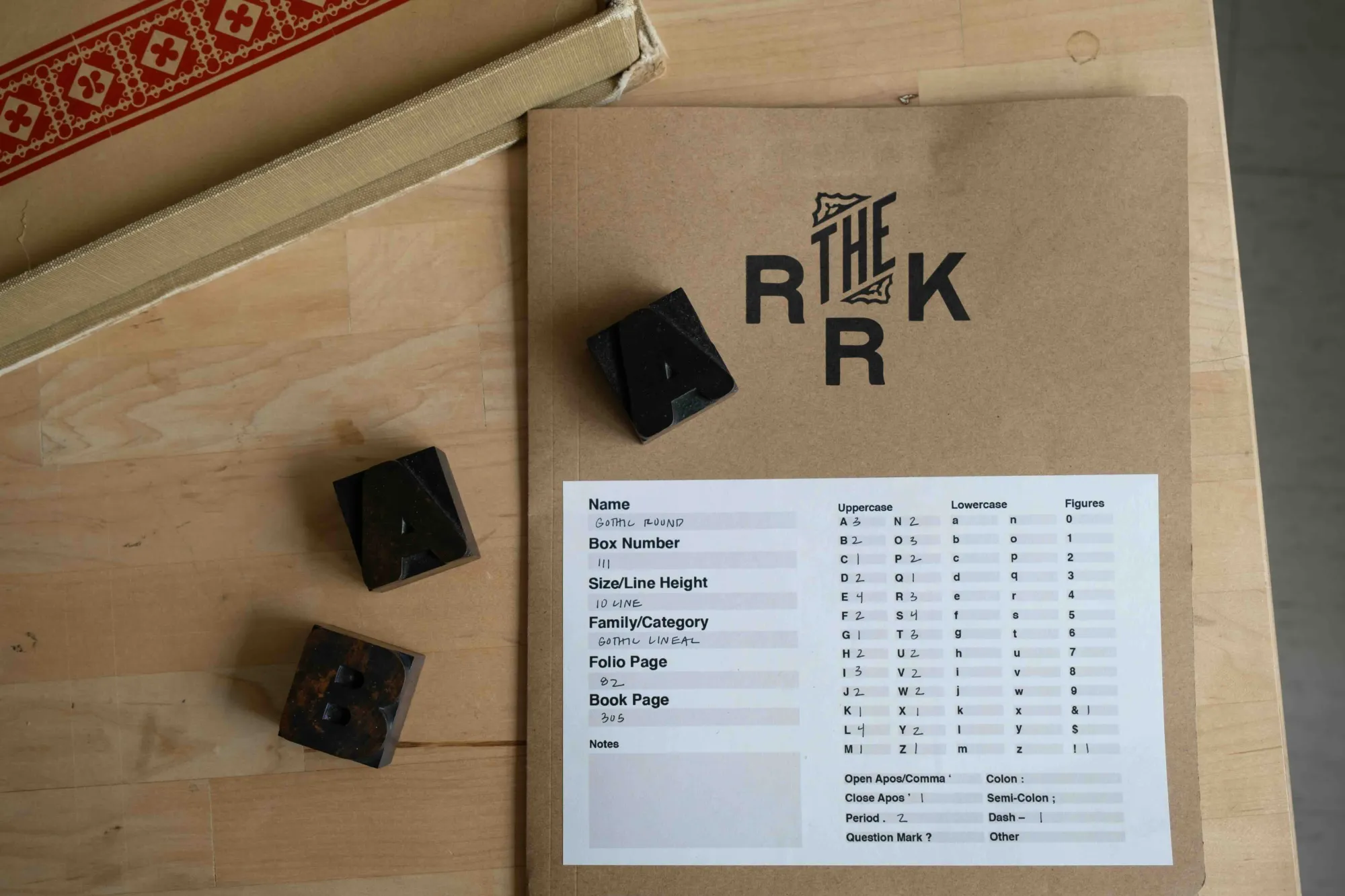 A close-up of a type specimen folder labeled “THE RRK” rests on a wooden table alongside three wood type blocks. The sheet inside details font name, size, and character availability, referencing the Gothic Round typeface. This documentation process is part of preserving and cataloging the Rob Roy Kelly American Wood Type Collection.