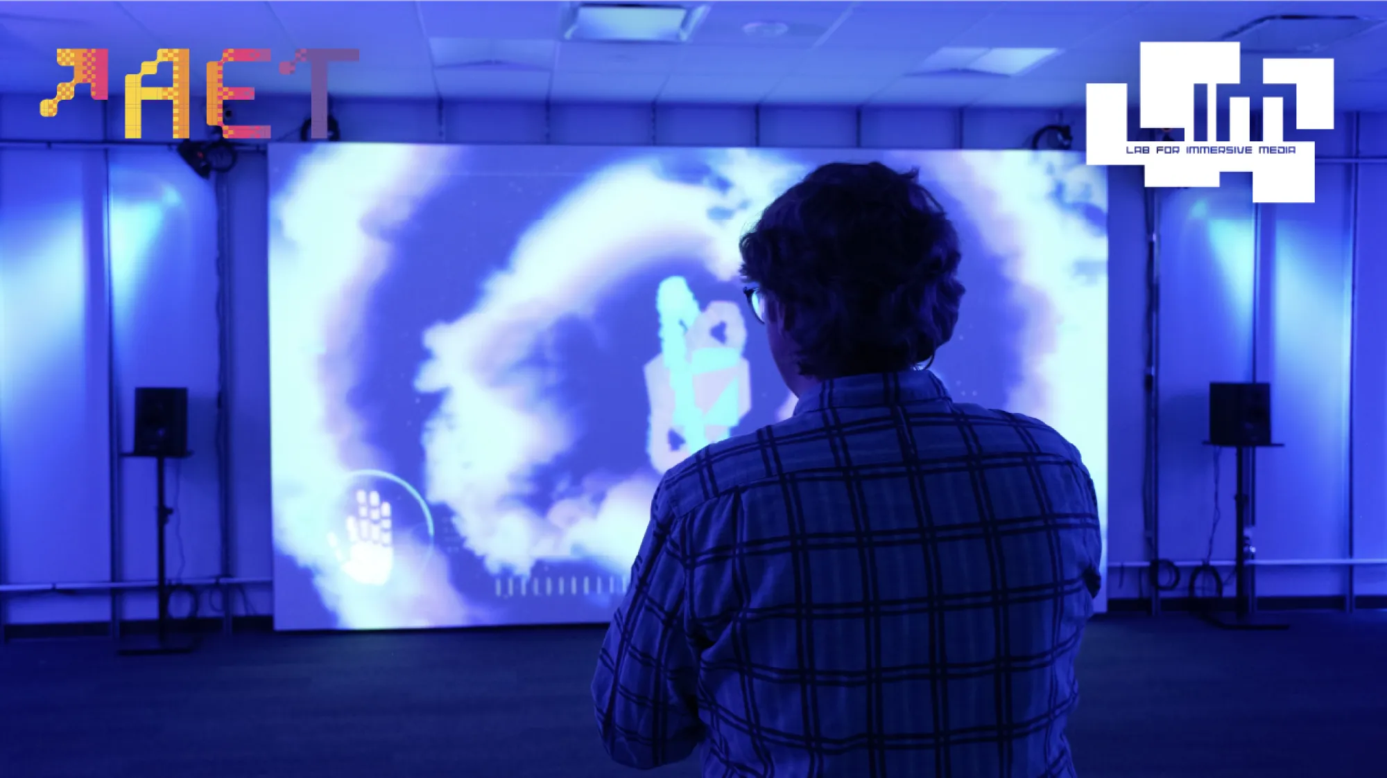 A person stands in a blue-lit immersive media room, facing a large projection screen displaying a vibrant digital interface with swirling abstract shapes and a glowing hand graphic. The logos for AET and the Lab for Immersive Media appear in the top-left and top-right corners, respectively.