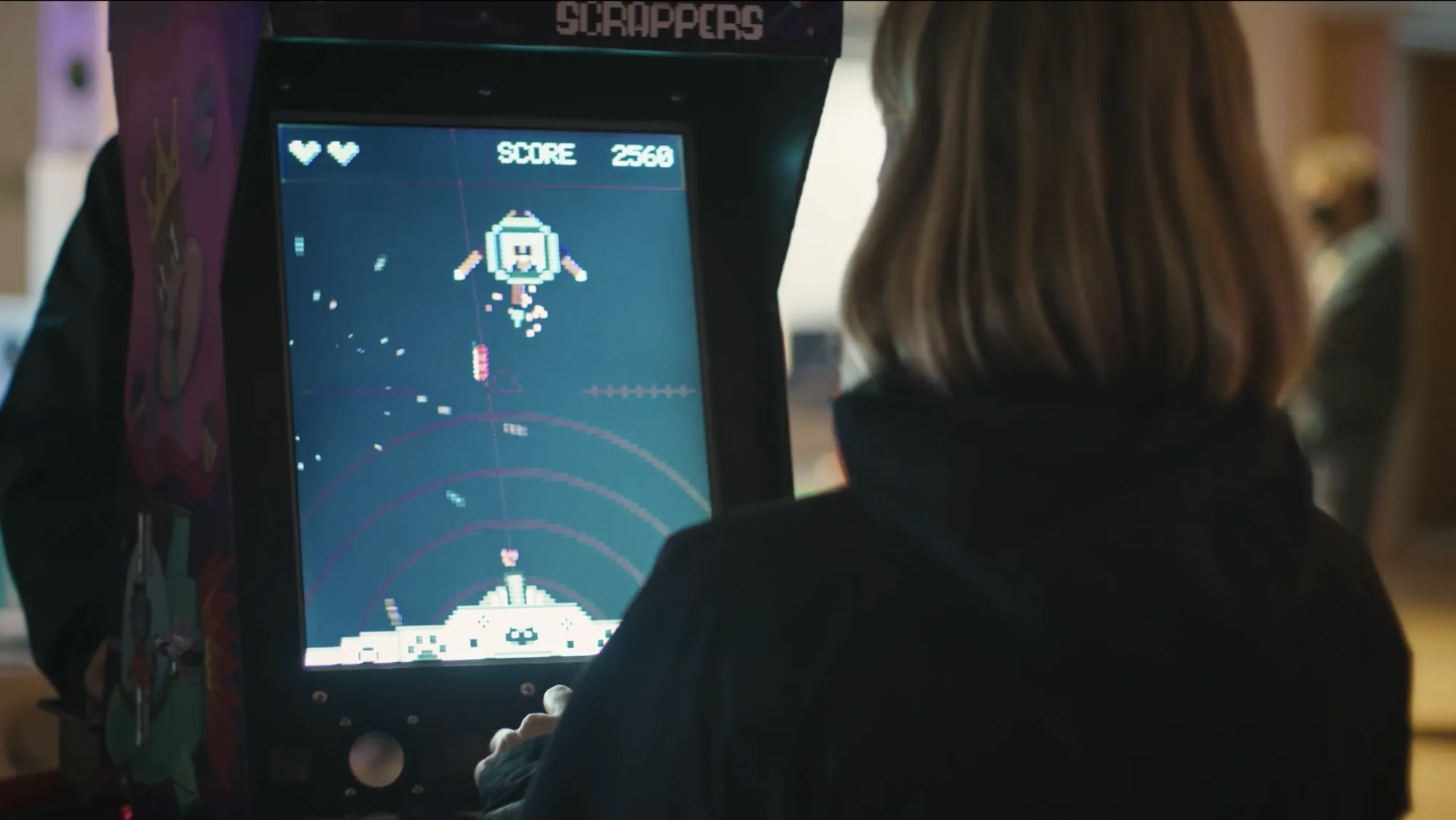 A person with shoulder-length hair plays a retro-style arcade game called “Scrappers.” The screen shows pixelated characters and a score of 2560, while neon visuals and joystick controls evoke a nostalgic yet playful atmosphere, representing AET’s emphasis on interactive media and game design.