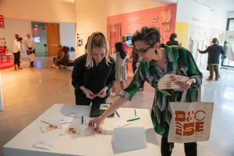 Graduate program faculty Kelcey Gray and Tamie Glass taking Becauseisms merch from a table