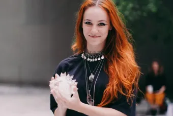 Person with long red hair and layered silver jewelry holds a translucent crystal sculpture outdoors, smiling warmly while looking directly at the camera. The background is softly blurred with greenery and another person in the distance.