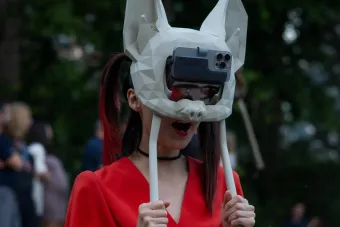 A person wearing a futuristic, geometric bat mask with a smartphone attached to the front, acting as a digital visor. The mask is white with angular facets, covering the entire head. The person, dressed in a striking red outfit, holds two white rods extending from the mask. Their mouth is open in an expressive pose. The background is blurred with an outdoor setting and a crowd of people, suggesting a live event or performance.