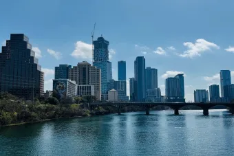 A wide view of the Austin skyline with high-rise buildings, the Colorado River in the foreground, and a bridge crossing the water.