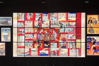 A large digital display wall showcasing a vibrant collage of Japanese woodblock prints. The central composition features a colorful, multi-panel artwork filled with scenes of traditional life, people in elaborate attire, and architectural elements, framed by red borders and Japanese text. Surrounding the center panel are smaller prints, depicting trains, Western visitors, marketplaces, parades, and scenic landscapes. The background is black, emphasizing the illuminated art pieces, which highlight a cultural