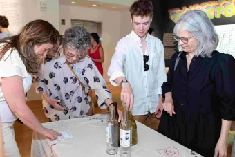 Four visitors gather around an embroidered tablecloth featuring stitched questions and designs, including “BE MINE” in a red heart. One person points to a label, while another places a bottle among others on the table. The colorful “TRAJECTORIES” title is visible on the exhibition wall behind them.