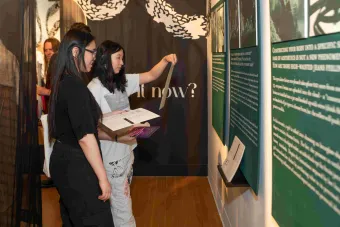 Two visitors stand side by side reading exhibition panels mounted on a gallery wall. One person holds up a printed sheet while the other reads from a program booklet. Large text in the background reads “it now?” beneath cloud-like graphics.