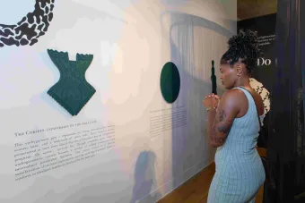 A person in a light blue ribbed dress stands with clasped hands, intently viewing a wall-mounted installation and text titled "The Corsets: Constraint of the Male Gaze." The display explores cultural narratives around corsets and gender expectations.