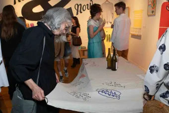 A woman with gray hair wearing a black dress closely examines the embroidered writing on a large textile laid out on a table. Around her, people mingle in a gallery filled with colorful artwork, and three wine bottles rest on the fabric.