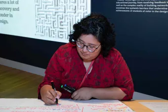 A person in a red floral top writes with colorful markers on a large sheet of paper covered in handwritten notes. Behind them, a black gallery wall displays text and a projected image of a maze.