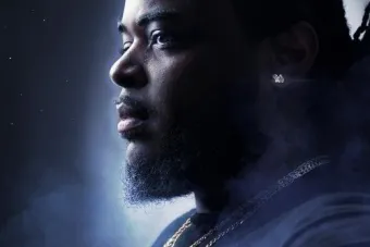 Portrait of Andrew Augustin looking to the side, wearing layered gold necklaces, with a misty background and soft lighting.