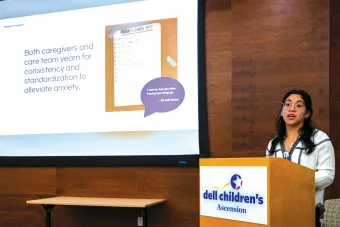 A presenter stands at a podium labeled “Dell Children’s Ascension,” delivering a talk in front of a projection screen. The slide reads, “Both caregivers and care team yearn for consistency and standardization to alleviate anxiety,” alongside an image of a checklist titled “DISCHARGE PREP” and a speech bubble quote from a caregiver. The presenter appears engaged and is mid-sentence, wearing glasses and a white top with dark embroidery. The room features wooden walls and a professional setting.