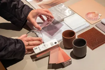 A person’s hands interact with a variety of material samples arranged neatly on a table. The samples include translucent acrylic sheets, color swatches, ceramic cups, fabric swatches, a shell, and textured tiles in neutral and earthy tones. The person holds a clear circular lens and a square plastic sheet, suggesting a material exploration or design planning activity.