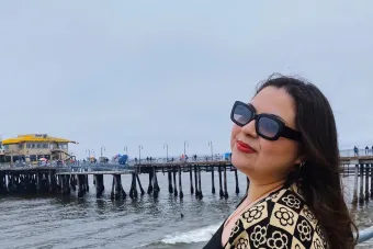 Xandria Hernandez wearing a patterned cream and black cardigan standing by the ocean with a wooden pier in the background under a cloudy sky.