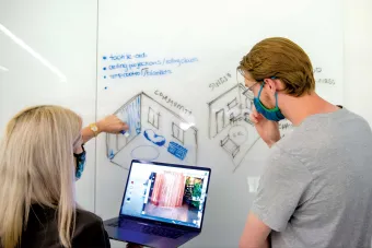 Two masked individuals collaborate in front of a whiteboard with hand-drawn room layouts labeled “COMMUNITY” and “SINGLE.” One person gestures at the drawings while the other looks on thoughtfully, holding a laptop displaying an image of a cylindrical structure. Blue handwritten notes mention tactile aids, ceiling projections, and temperature control.