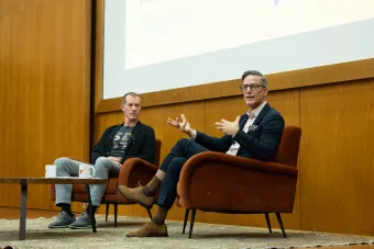 Two speakers sit on a stage in rust-colored chairs during a moderated talk. One gestures while speaking, and the other listens attentively. A projected screen behind them displays the School of Design and Creative Technologies logo. A table in front of them holds two mugs with the Longhorn logo.