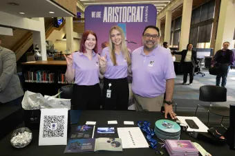 Three Aristocrat Gaming representatives pose at a recruiting table with marketing materials and giveaways inside a campus library space.