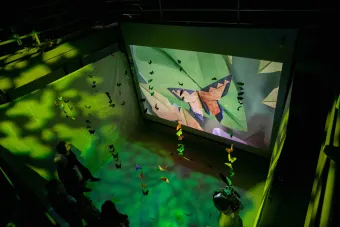 Overhead view of an immersive butterfly-themed installation featuring hanging origami butterflies, colorful green lighting effects, and a large screen projection of a folded paper butterfly.