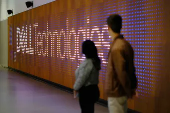 Two students stand in front of an interactive digital wall display featuring the Dell Technologies logo illuminated in white and purple lights.