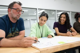 Three individuals sit at a table in a bright, modern classroom, collaborating on a brainstorming activity. One person draws on a large sheet of paper while others look on, smiling and engaged. Sticky notes and sketching materials are scattered across the table.