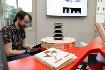 Student examines a tablet while seated beside a colorful, sculptural display featuring stacked discs, patterned graphics, and printed booklets. A digital screen with design work is visible in the background.