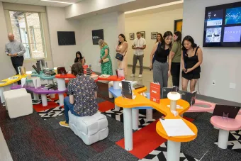 Visitors explore a design exhibition featuring colorful tables displaying books, lights, and sculptural objects. Attendees observe and interact with the work in a well-lit room with modern decor and design-focused posters on the walls.