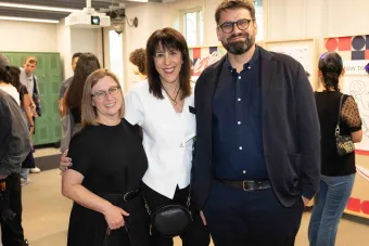 Three adults stand smiling for a photo at a design exhibition opening. They are surrounded by other attendees in a bright, modern space with colorful artwork on the walls and green lockers in the background.