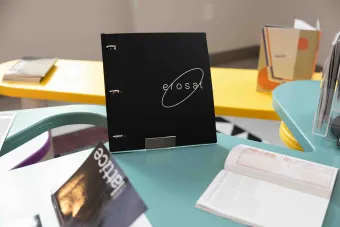 Black binder with white text reading “erosat” on display atop a teal table, surrounded by other printed student publications at a design showcase.