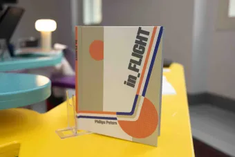 Close-up of a publication titled “in.FLIGHT” by Phillips Peters, displayed upright on a yellow table. The cover features retro-inspired graphics in orange, blue, and beige tones.