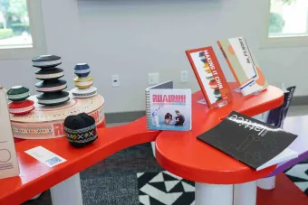 Colorful table display featuring Chin cultural artifacts and design work, including a stacked bowl sculpture, embroidered hat, and books such as “Rual Dih! Rule Book,” “Making It Chin,” “in.FLIGHT,” and a black-covered issue of “latitce.”