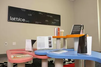 Exhibition view of the Lattice MFA in Design showcase featuring colorful modular tables displaying student publications like “Erosat,” “in.FLIGHT,” and “lattice.” A large black wall sign lists the exhibit titles and designers.
